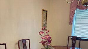 Ein Esstisch mit floraler Dekoration, umgeben von Stühlen, in einem Raum mit einer Kristallleuchtre und einem fenster mit Jalousien.