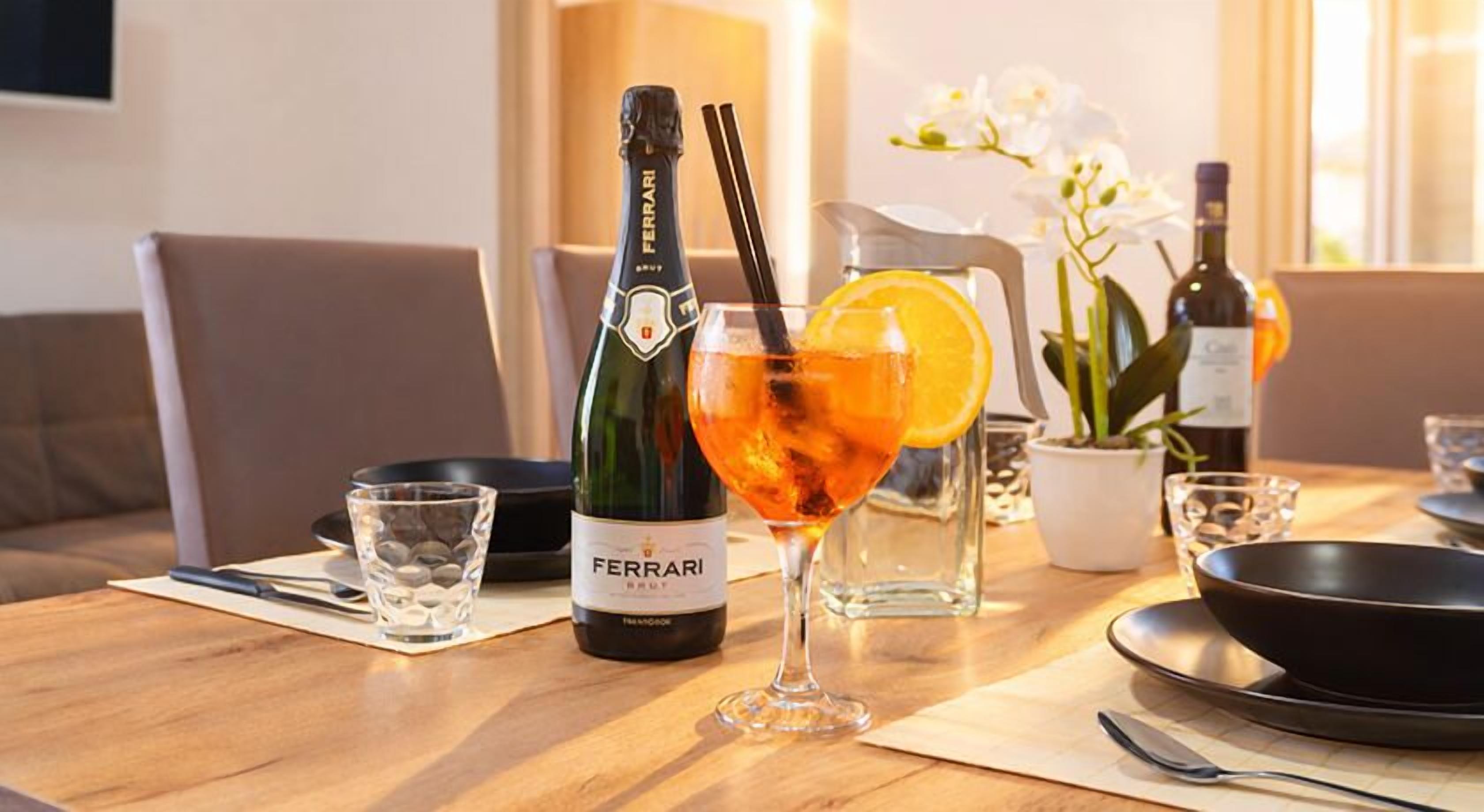 Una mesa de comedor con una botella de vino Ferrari, una copa de vino y un cóctel con una rodaja de naranja.
