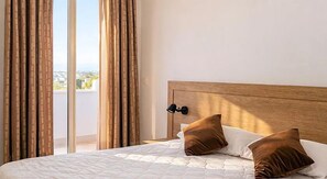 A hotel room with a bed, two pillows, a nightstand, a lamp, and a view through a window with curtains. - Kalispera Residence  (Scalea)
