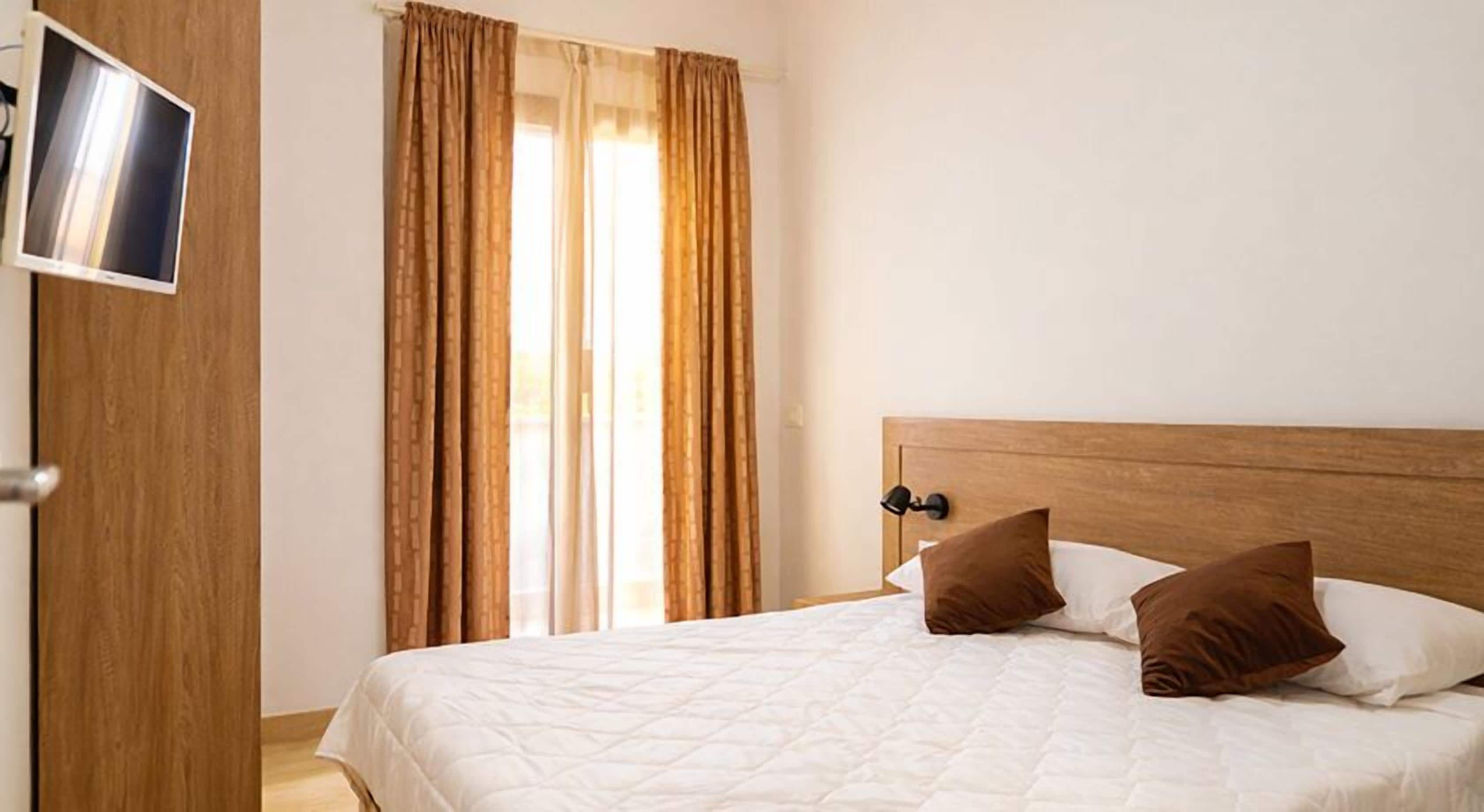 Camera d'albergo con un letto, una testiera in legno, due cuscini, una TV montata su una porta di legno e una finestra con tende.