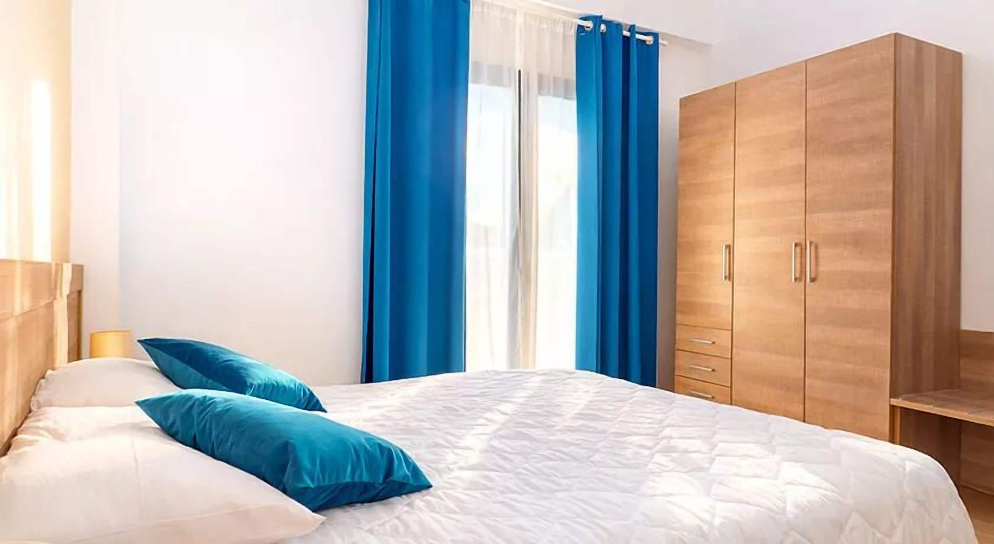 A bedroom with a bed, blue curtains, a wooden wardrobe, and a bedside lamp.