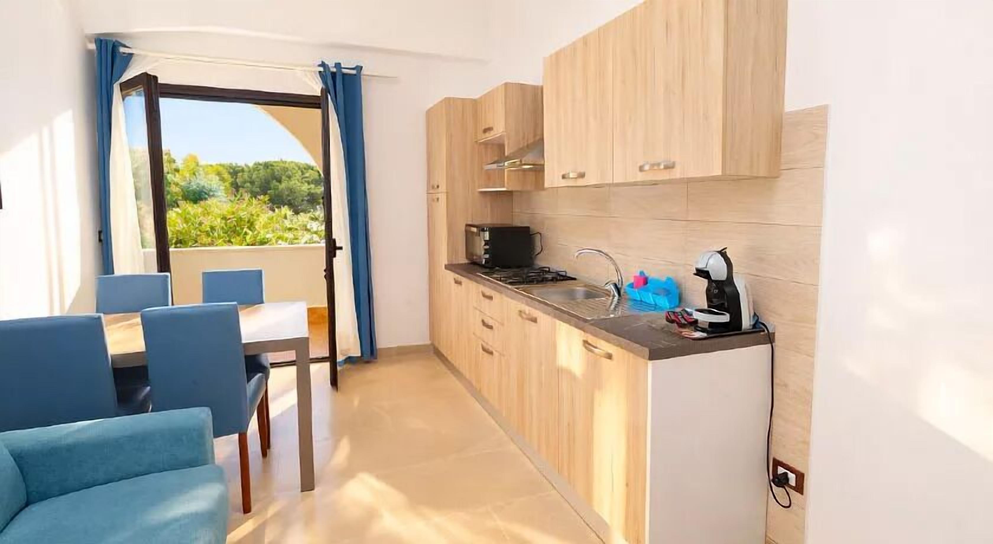 A compact kitchen with wooden cabinets, a gas stove, and a coffee machine. There is a dining area with blue chairs and a table.