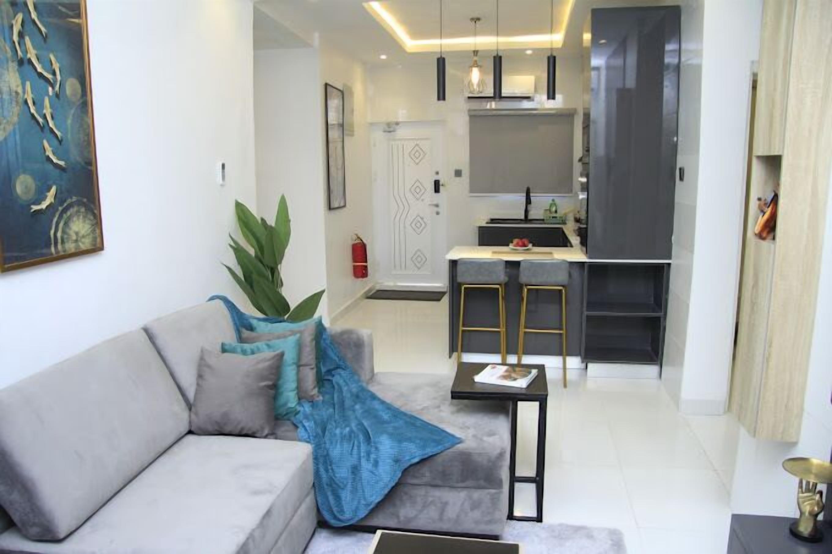 Una sala de estar moderna con un sofá gris, una mesa de centro y una planta. Se observa una cocina con un refrigerador y un lavamanos al fondo.