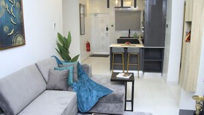 Ein modernes Wohnzimmer mit einem grauen Sofa, einem Couchtisch und einer Pflanze. Im Hintergrund befindet sich eine Küchenzeile mit Kühlschrank und Spüle.
