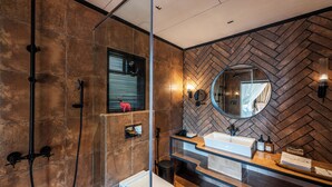 A modern bathroom with a herringbone-patterned wall, a round mirror, a glass shower enclosure, and a dual sink vanity. - Canary Islands Resort & Spa (Paud)