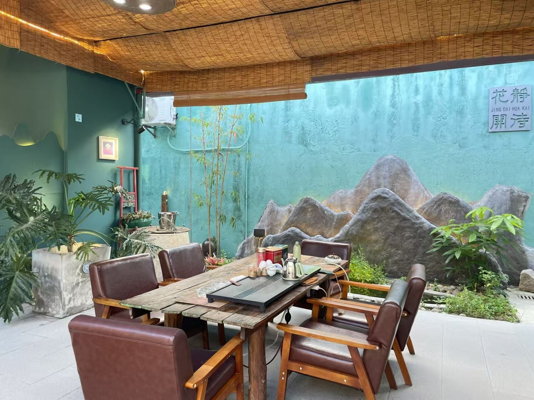 Comedor con una mesa de madera rústica y sillas de cuero, rodeado de plantas en macetas y un mural de rocas y follaje.