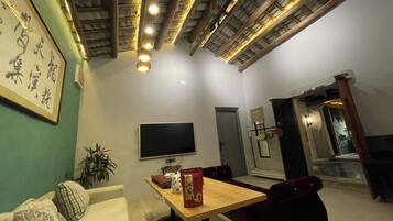 A living room with a sofa, a dining table, a TV, and a ceiling with exposed beams and lights.