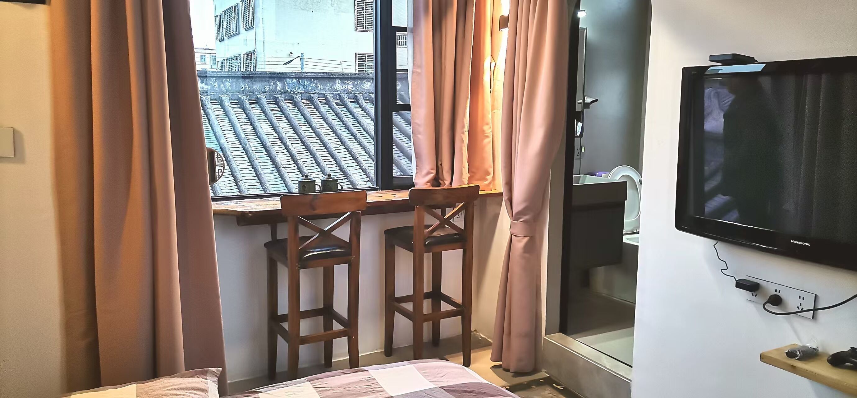 Une pièce dotée d’une fenêtre donnant sur un toit traditionnel recouvert de tuiles, d’un téléviseur fixé au mur et d’une petite table en bois avec deux chaises.