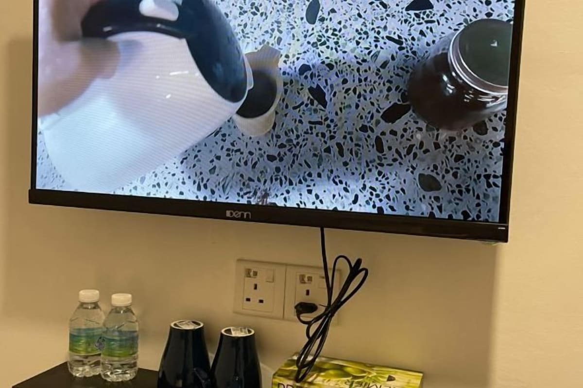 A television mounted on a wall with a power outlet and cables below it. There are two water bottles and a black object on the table in front of the TV.