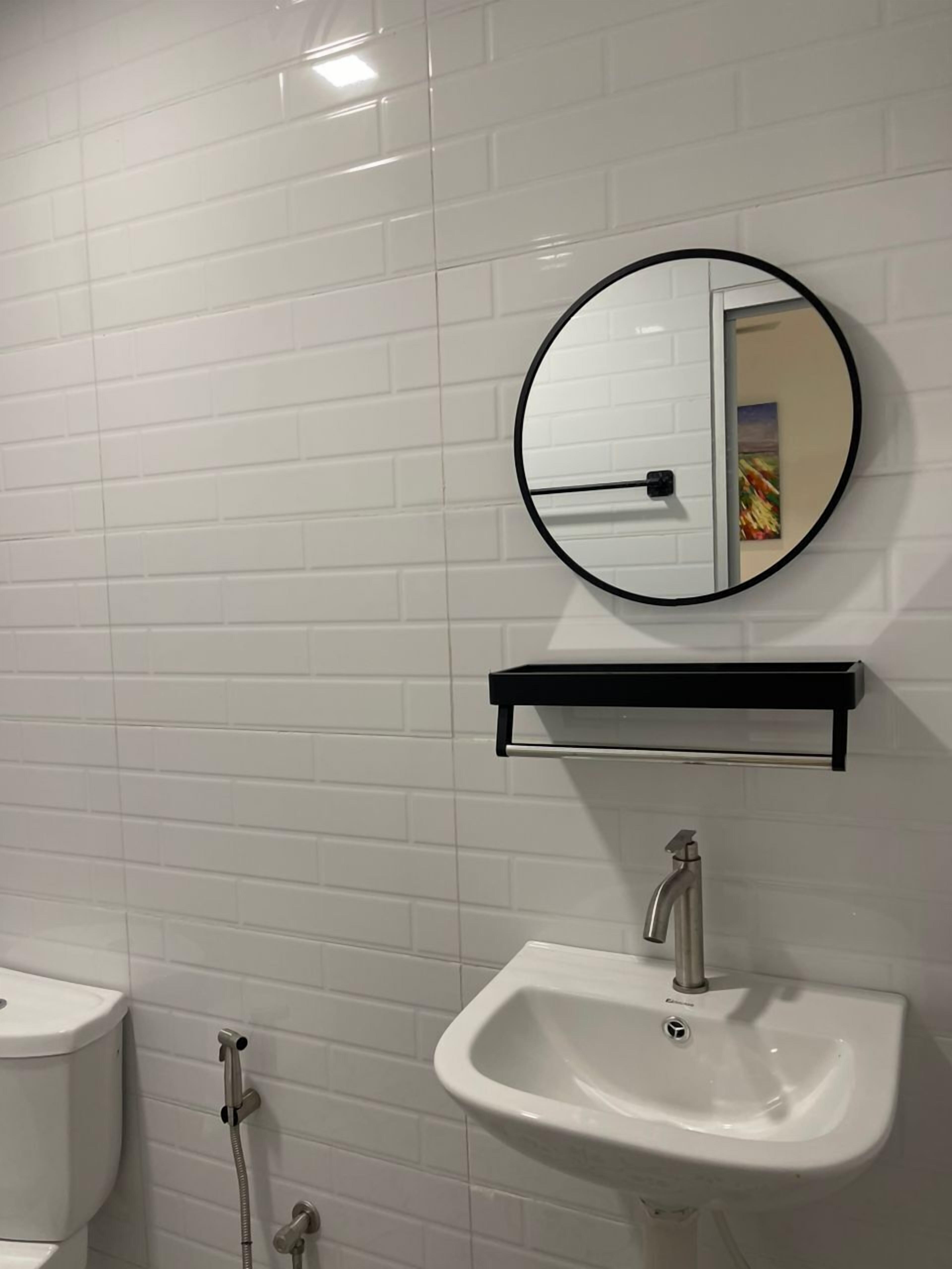 Un baño con paredes de azulejos blancos, un lavamanos blanco, un espejo redondo y un estante negro.
