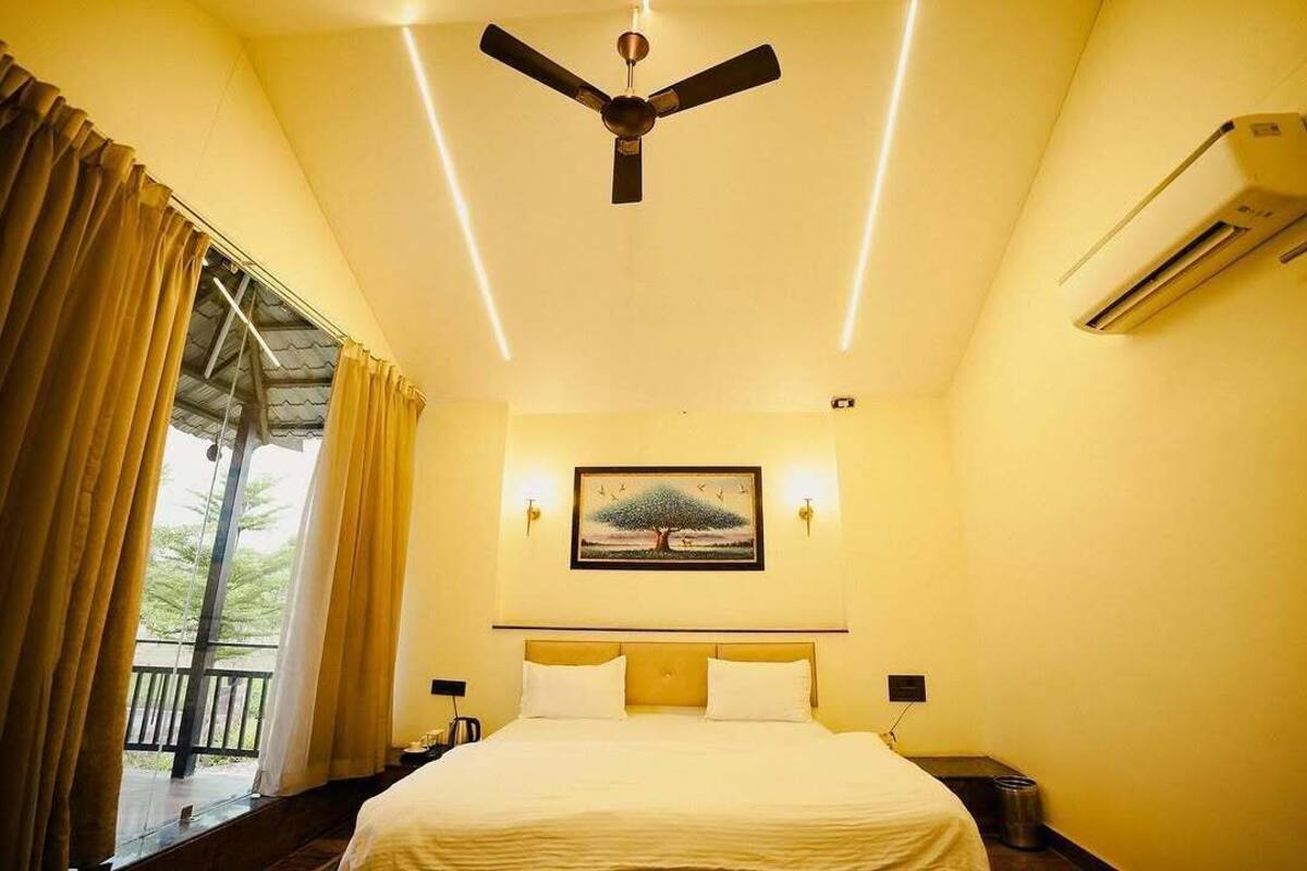 A hotel room with a bed, a ceiling fan, an air conditioning unit, a painting on the wall, and a window with curtains.