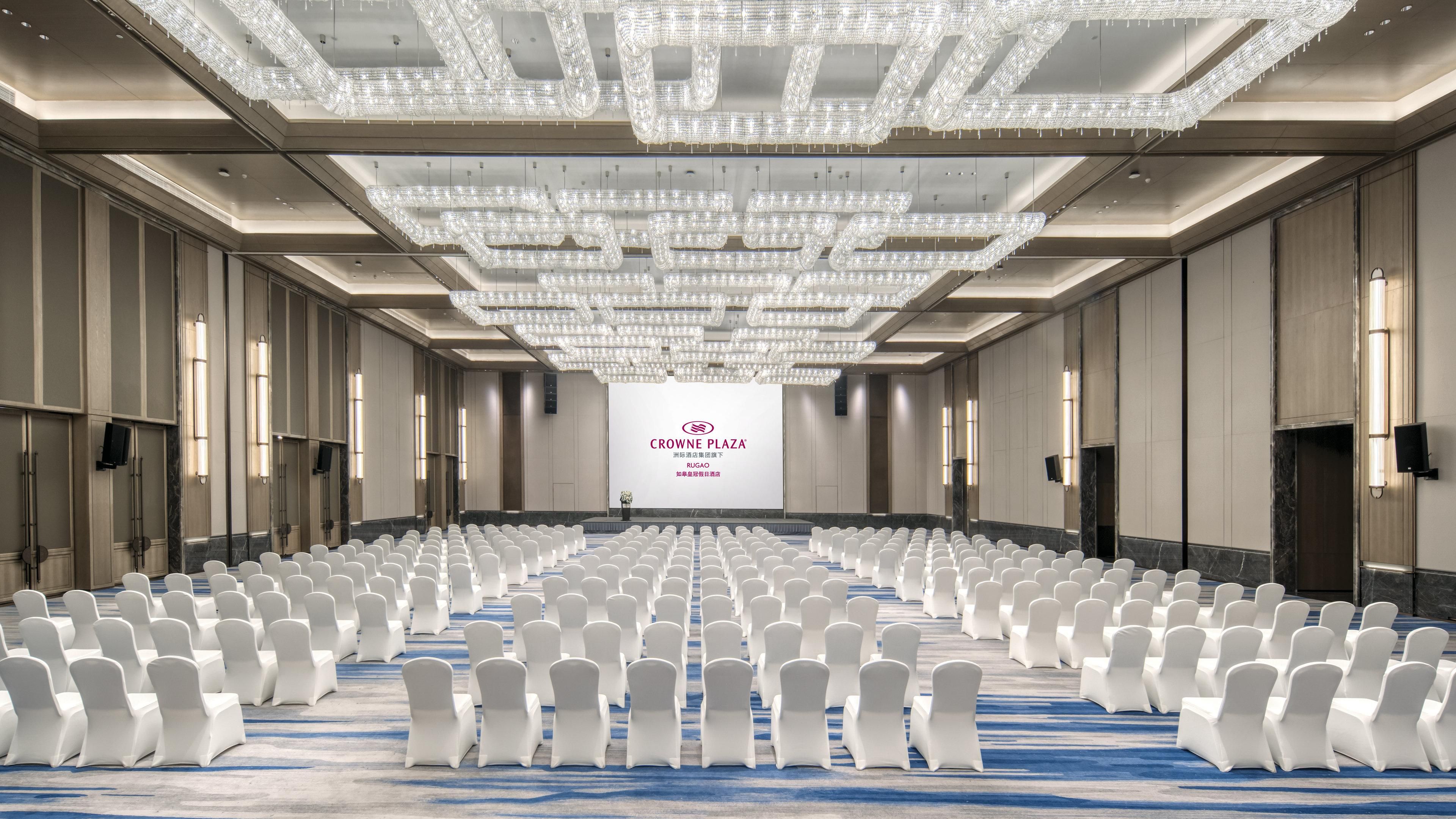 Un salón de conferencias con filas de sillas blancas, una pantalla grande mostrando 'Crowne Plaza RUGAO' y una araña.