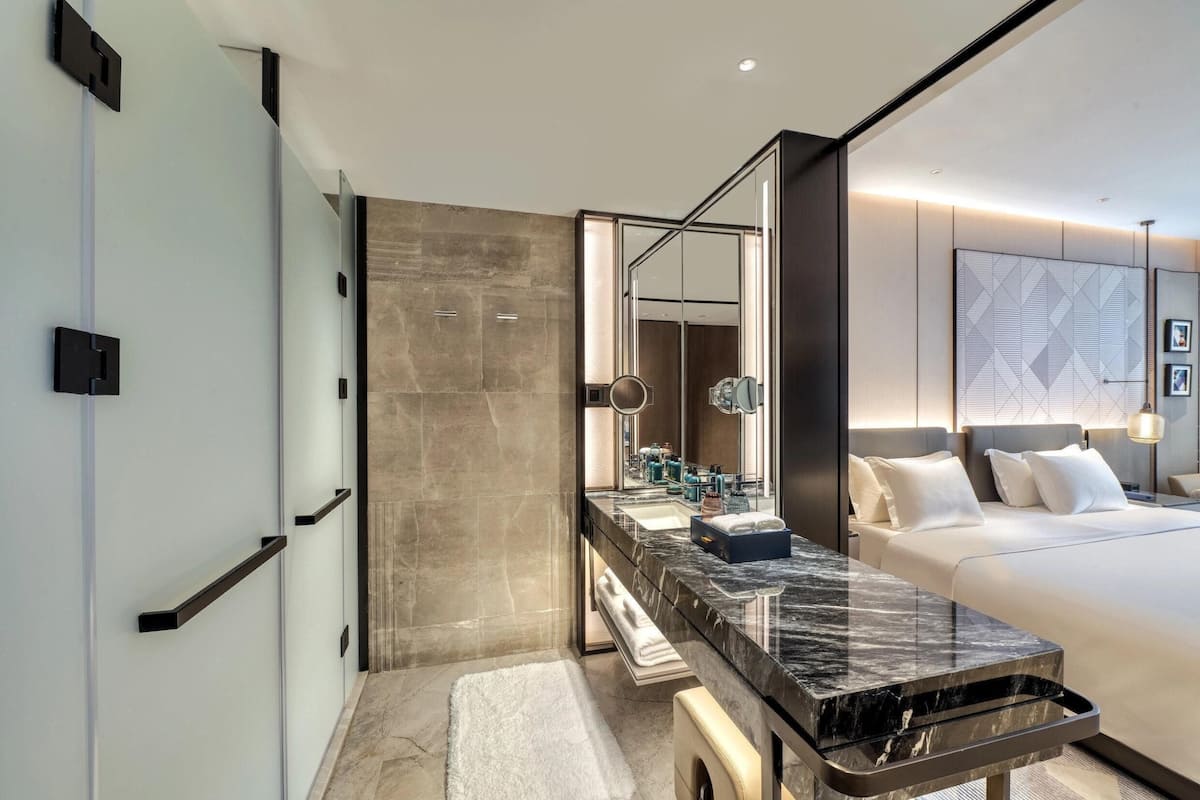 A modern hotel room with a marble bathroom vanity, a large bed, and a wall-mounted mirror.