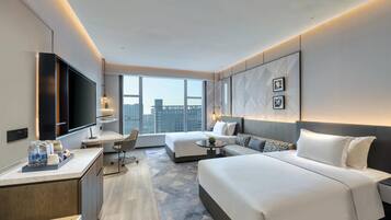 A modern hotel room with a large bed, a desk with a flat-screen TV, a seating area with a sofa and armchairs, and a city view through large windows.