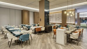 Un restaurant moderne doté de nombreuses tables et chaises, d’un bar central et d’une cuisine visible au fond de la salle.