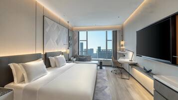 A modern hotel room with a large bed, a desk, a chair, a TV, and a city view.