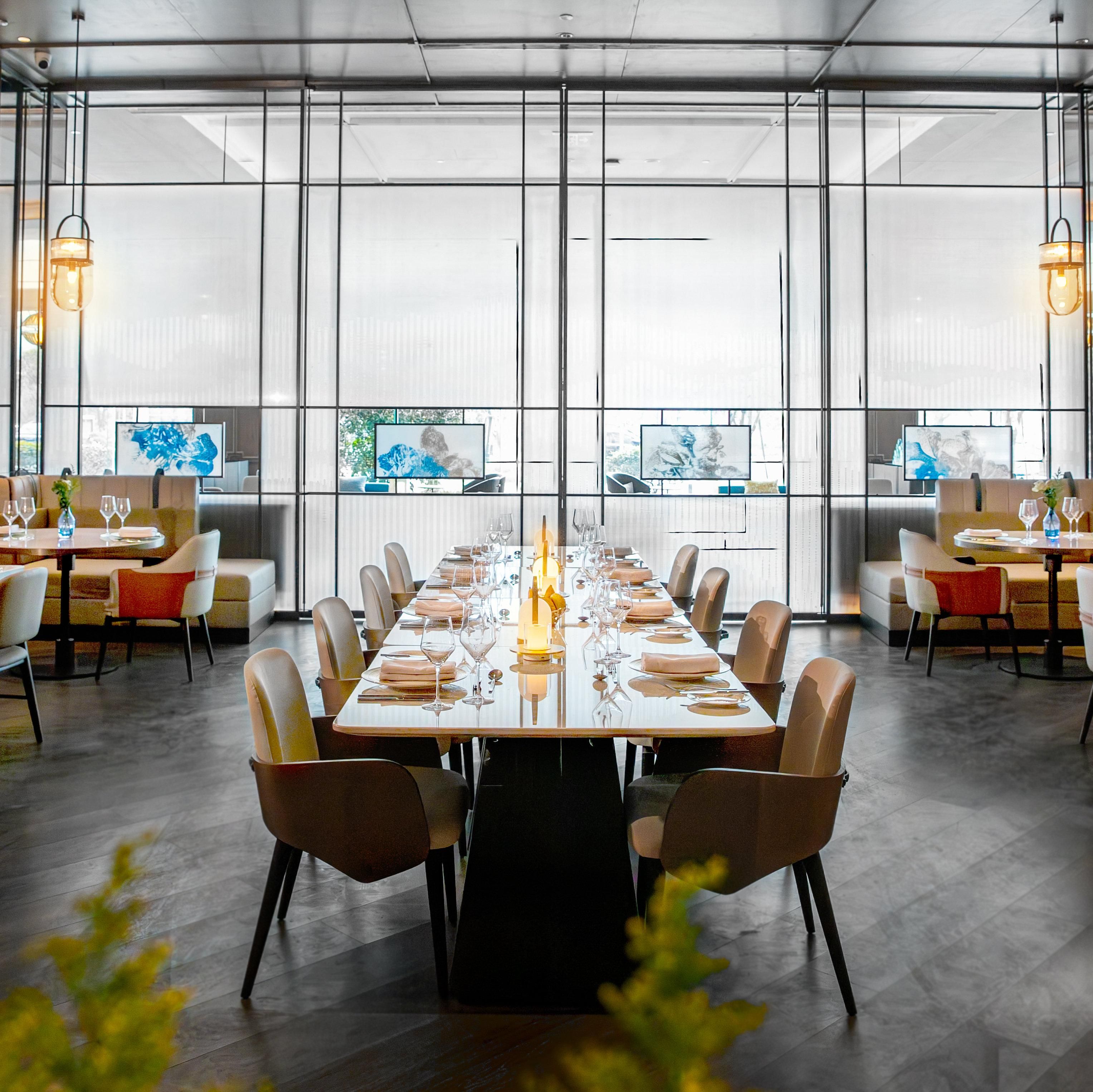 Un comedor moderno con una larga mesa dispuesta para una comida, ventanas amplias y obras de arte en las paredes.