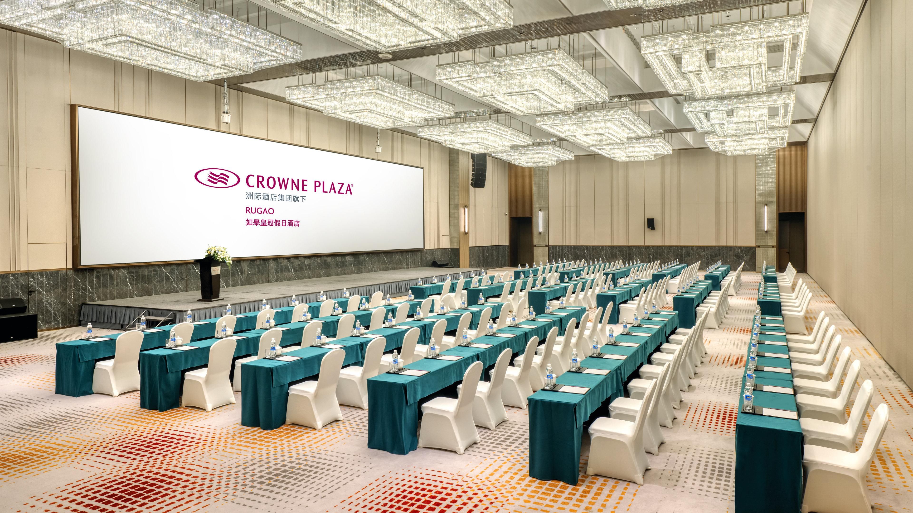A conference room with rows of tables and chairs, a large screen displaying text, and a stage with a podium.