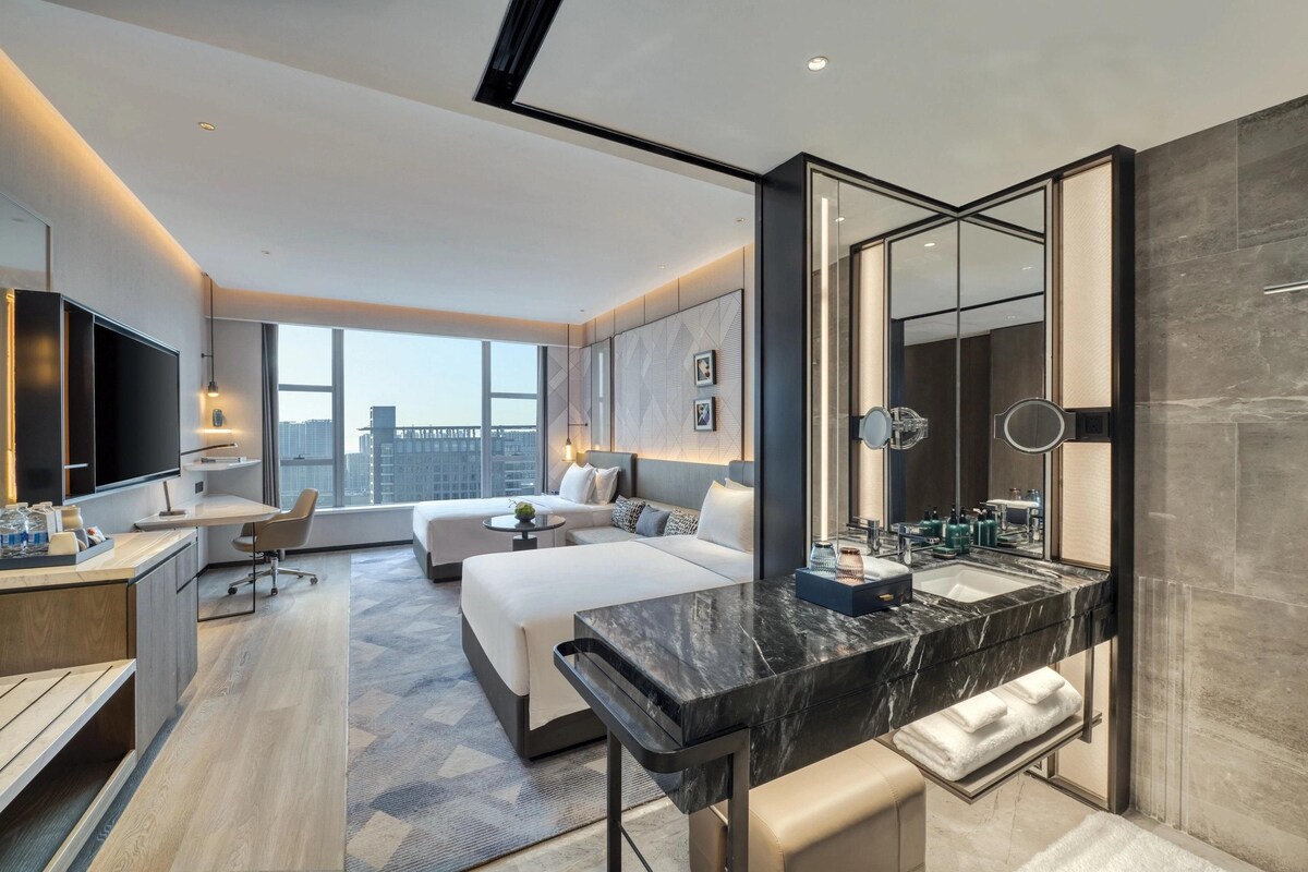 Une chambre d’hôtel moderne dotée d’un grand lit, d’un bureau, d’un coin salon, d’un grand miroir et d’une salle de bain avec un comptoir en marbre.