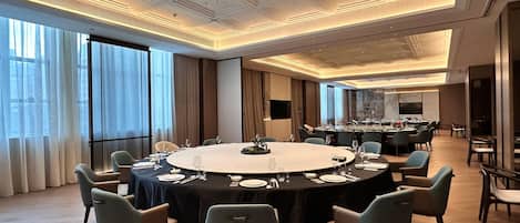 Uma sala de conferências com uma grande mesa oval, rodeada por cadeiras, num ambiente moderno de hotel com janelas amplas e pé-alto.