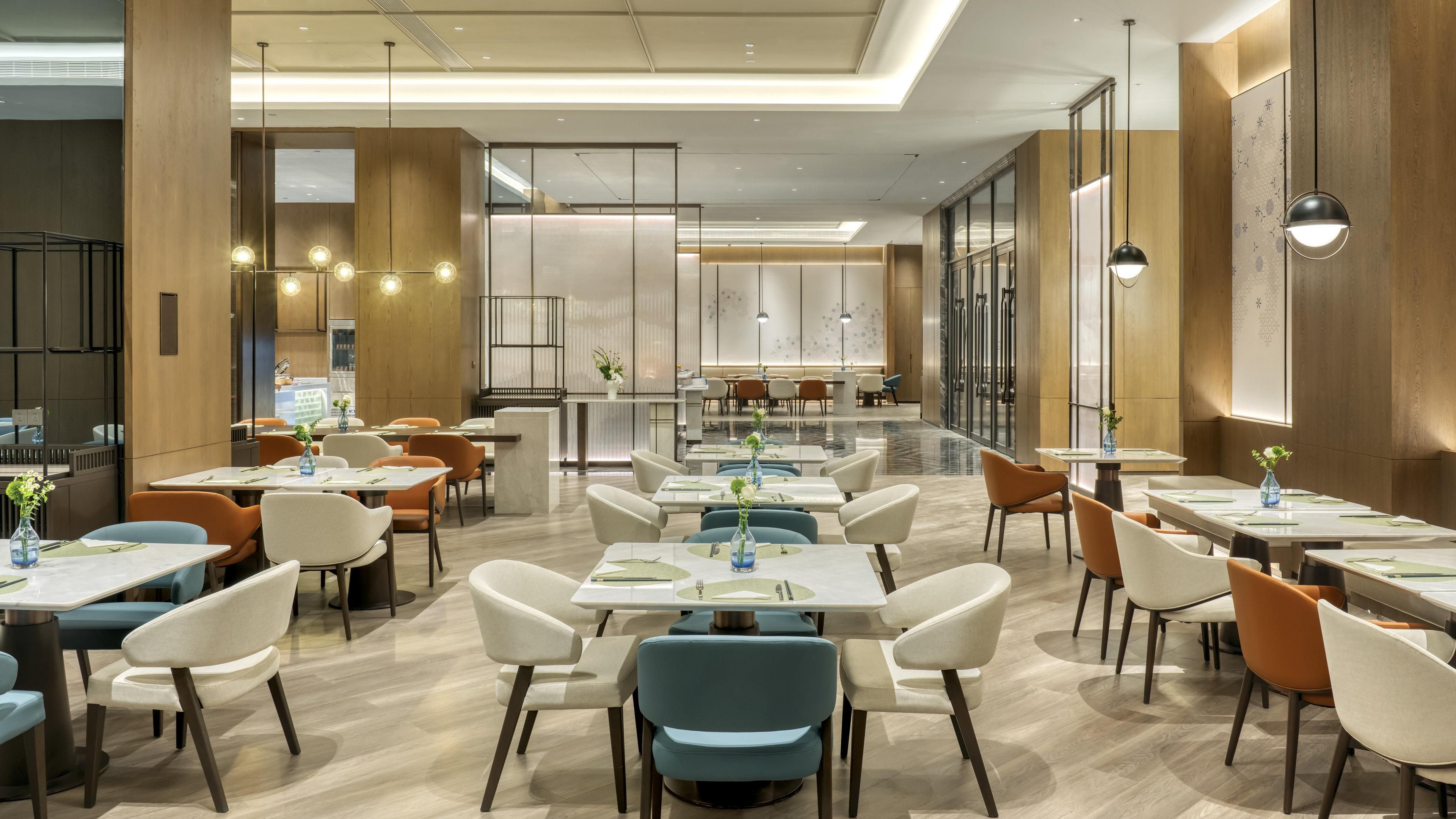Un restaurante moderno con un diseño espacioso, que cuenta con varias mesas y sillas, una zona de bar central y grandes ventanales que permiten la entrada de luz natural.