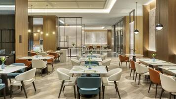 A modern restaurant with a spacious layout, featuring multiple dining tables and chairs, a central bar area, and large windows providing natural light.