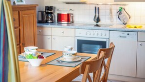 Una cocina con armarios de madera, una cafetera, una olla roja y una mesa de comedor con un tazón de fruta y tazas.