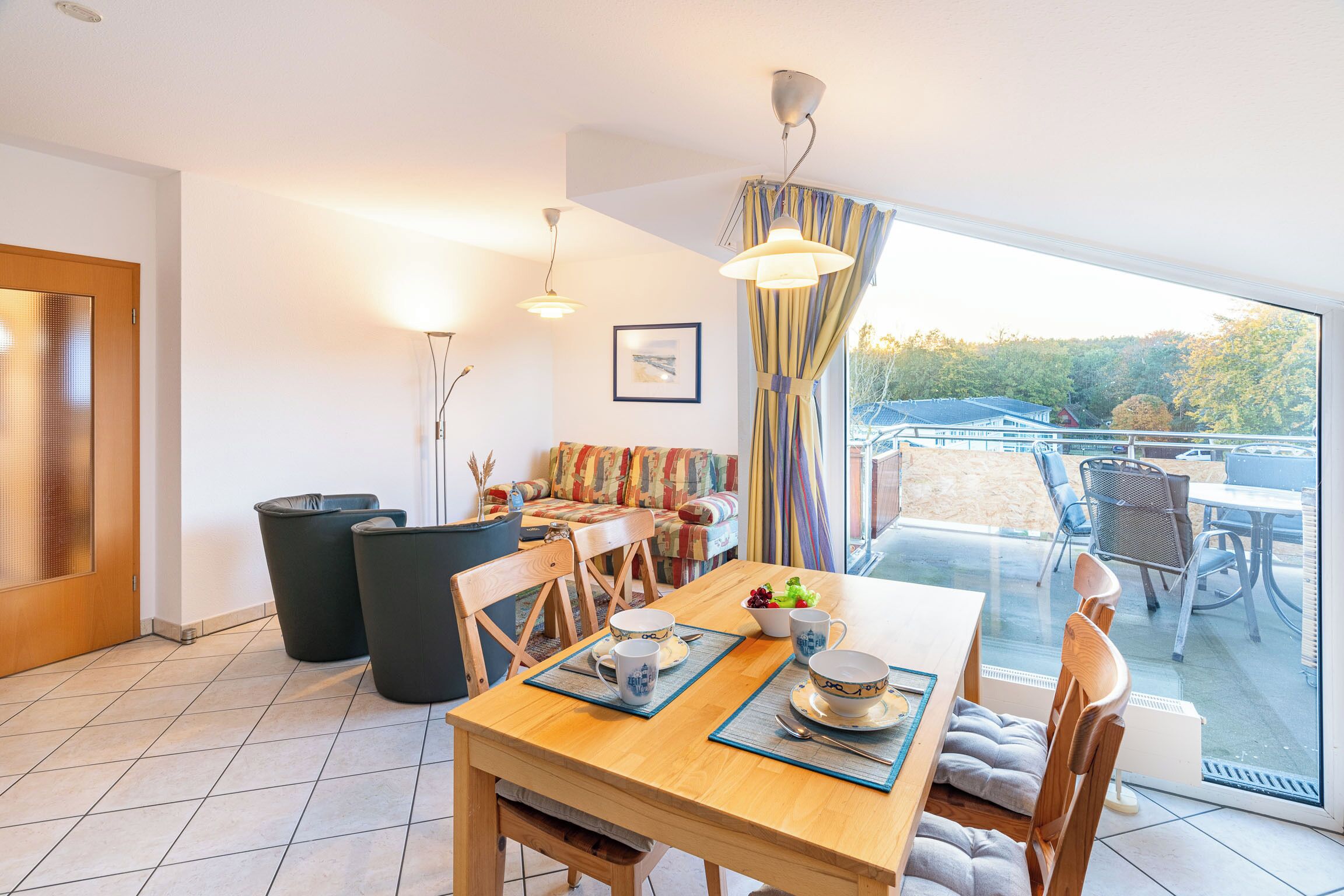 A dining area with a wooden table set for four, chairs, a buffet table with food, and a view of the outdoors through large windows.