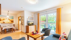 Ein Wohnzimmer mit Holztisch, Sofa, Sesseln, Fernseher und Blick ins Freie.