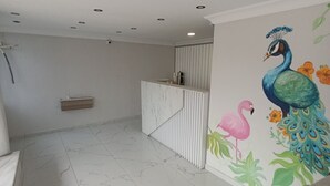 En rummelig stue med lyse marmorgulv, et hvidt receptionistskranke og et farverigt vægmaleri af påfugl og flamingo.