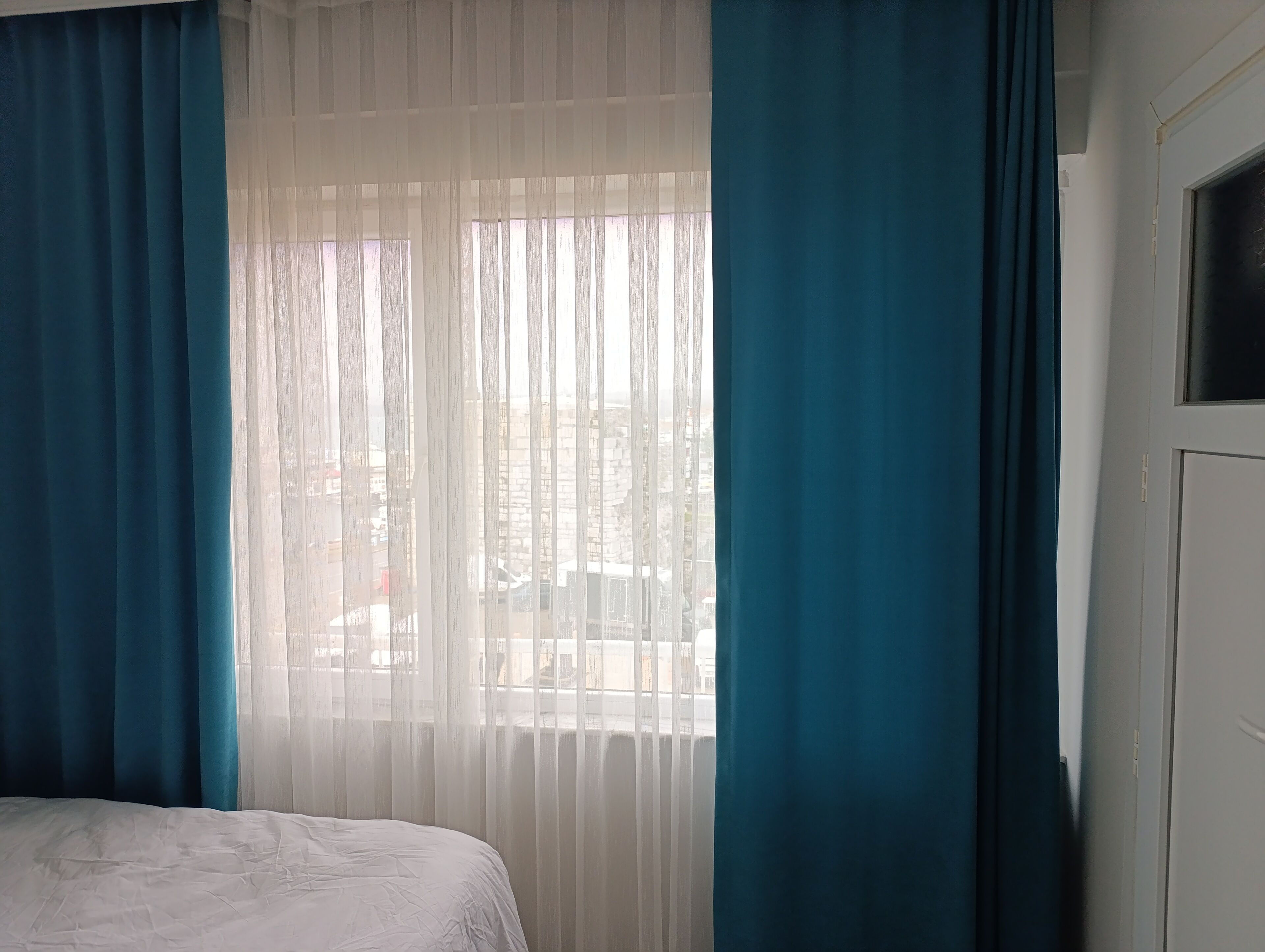 Ein Schlafzimmer mit Bett, einem Fenster mit durchsichtigen Vorhängen und Blick auf die Straße.