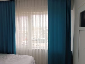 Un dormitorio con cama, ventana con cortinas translúcidas y vista a la calle.