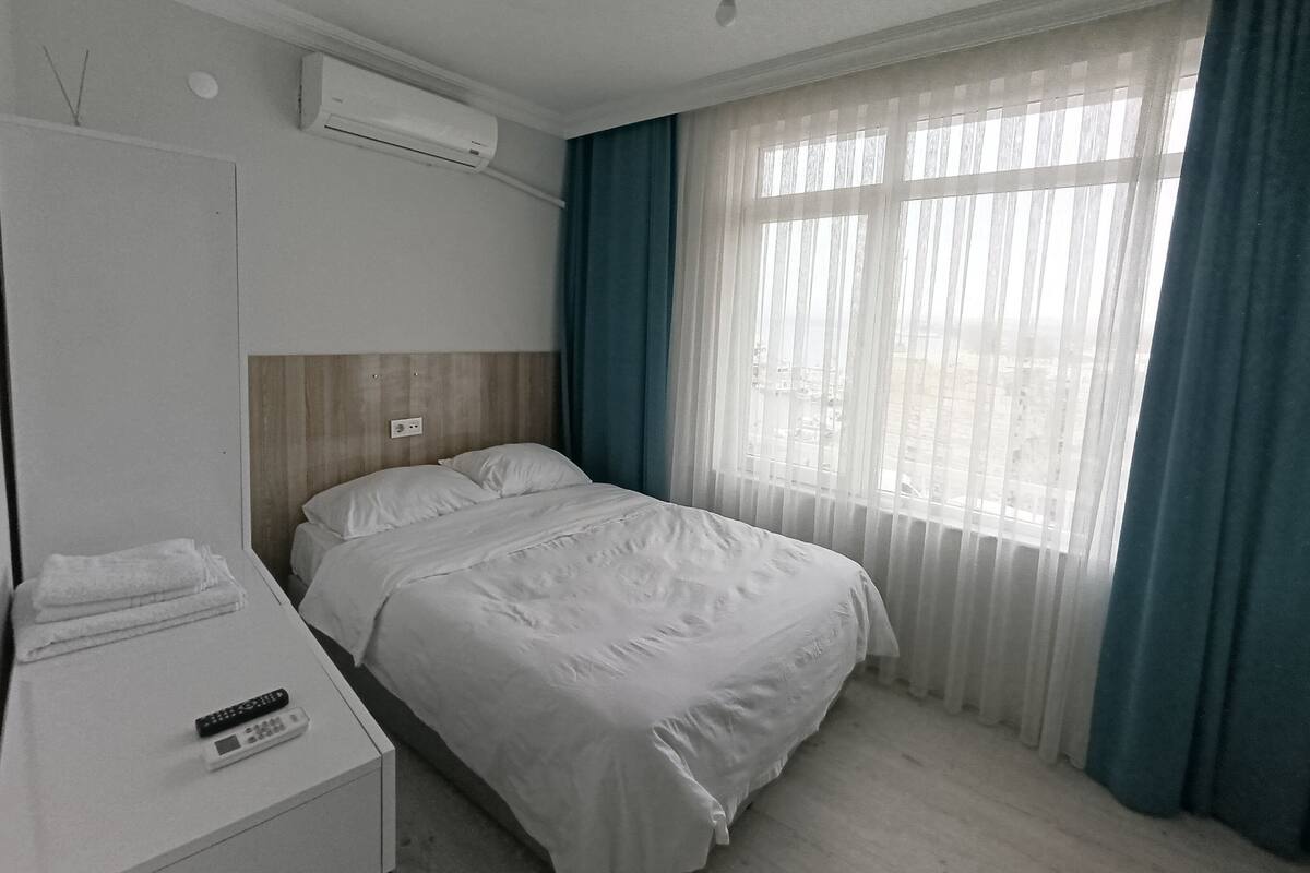 A hotel room with a bed, a bedside table with a remote control, an air conditioning unit, and a window with curtains.