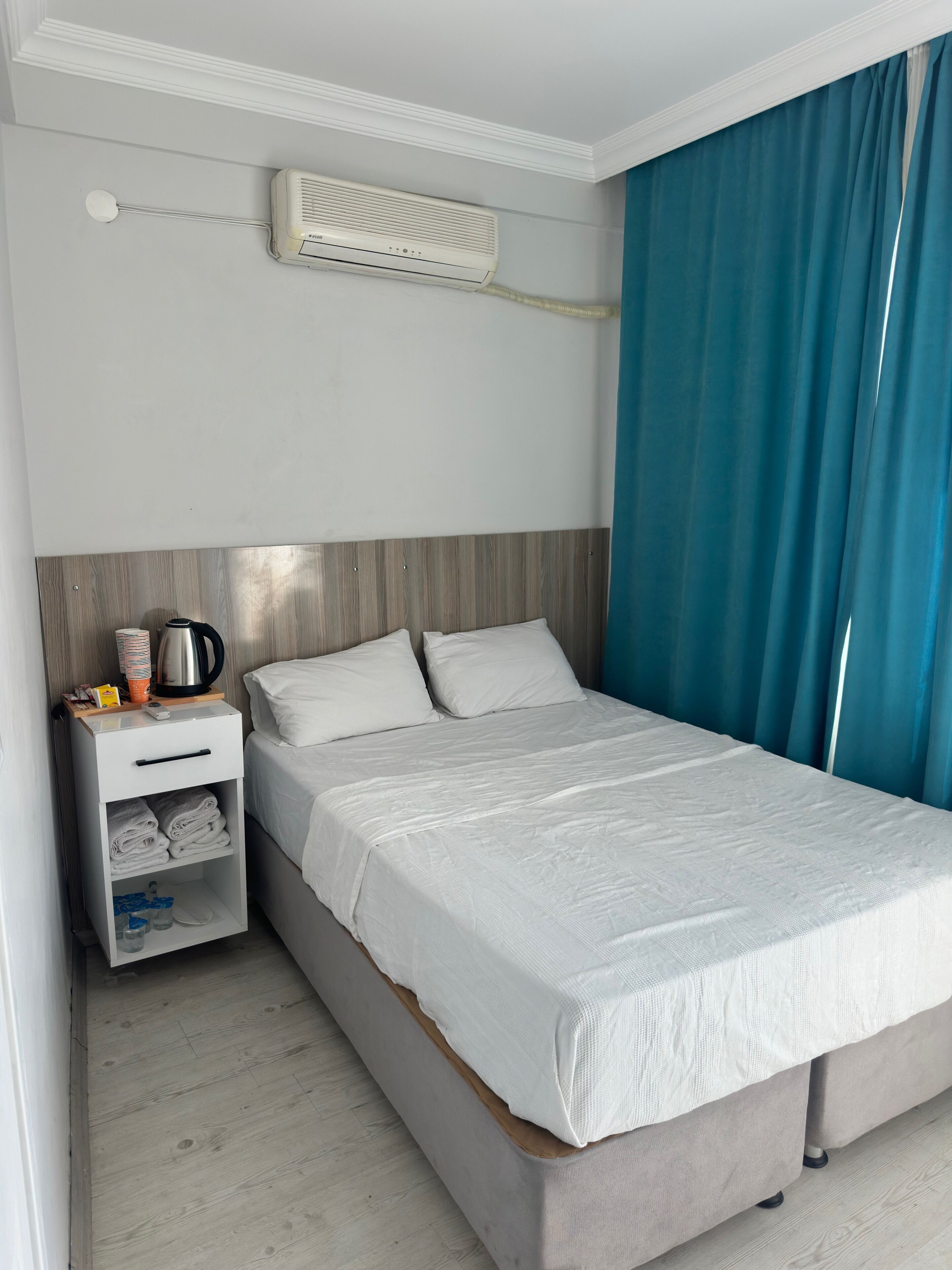 A hotel room with a bed, a nightstand, a heater, and a window with curtains.