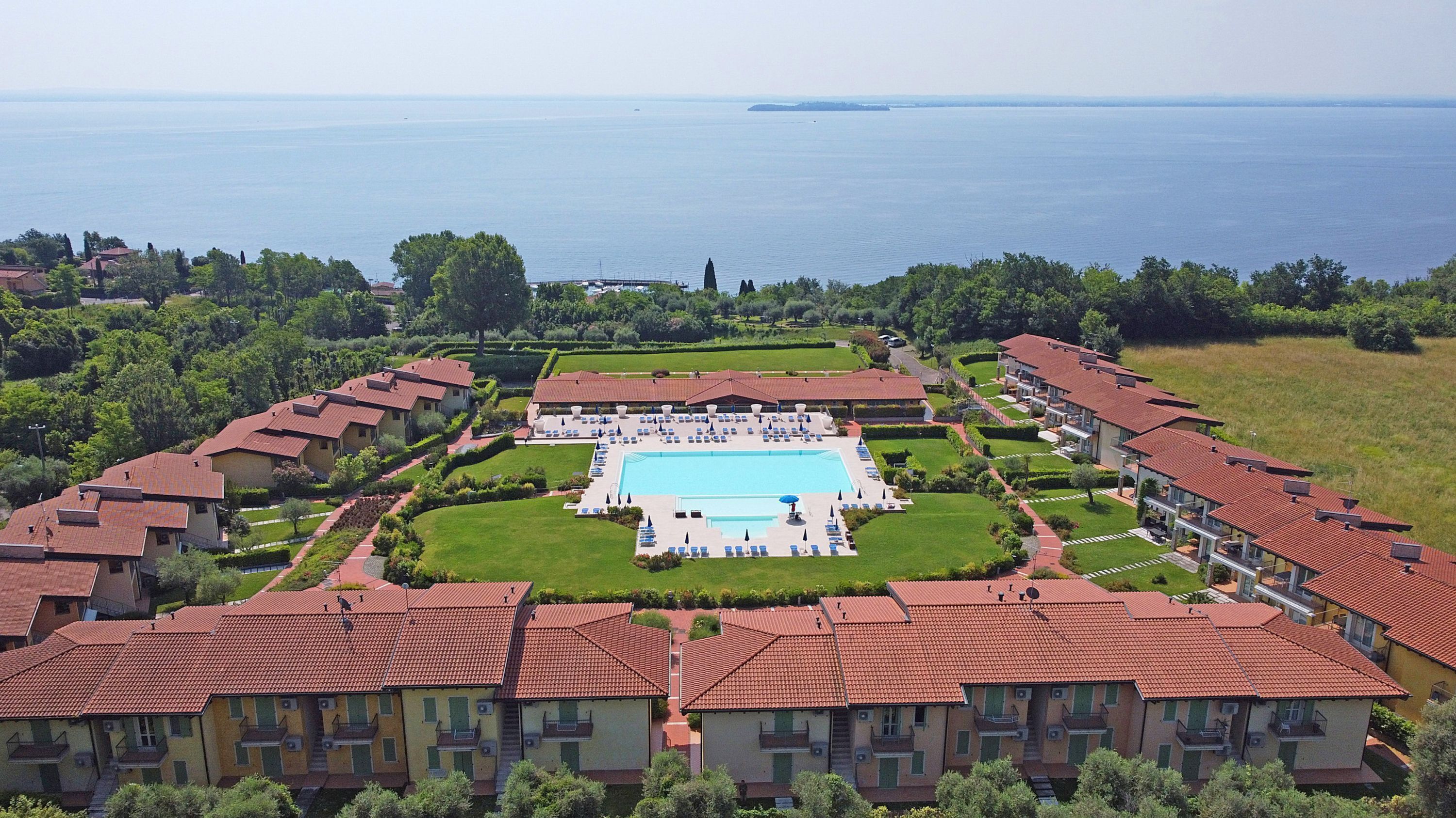 A resort with a central swimming pool, multiple guest rooms, and a view of the sea.