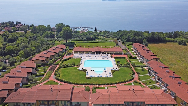 Luchtbeeld van een resort met een centraal zwembad, omringd door gelijkmatig ontworpen villa's met rode pannendaken, en een jachthaven op de achtergrond.