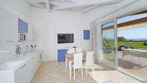 Een moderne keuken met witte kasten, een magnetron en een televisie. Er is een eethoek met een tafel en stoelen, en een schuifpui die leidt naar een terras met uitzicht op het landschap.