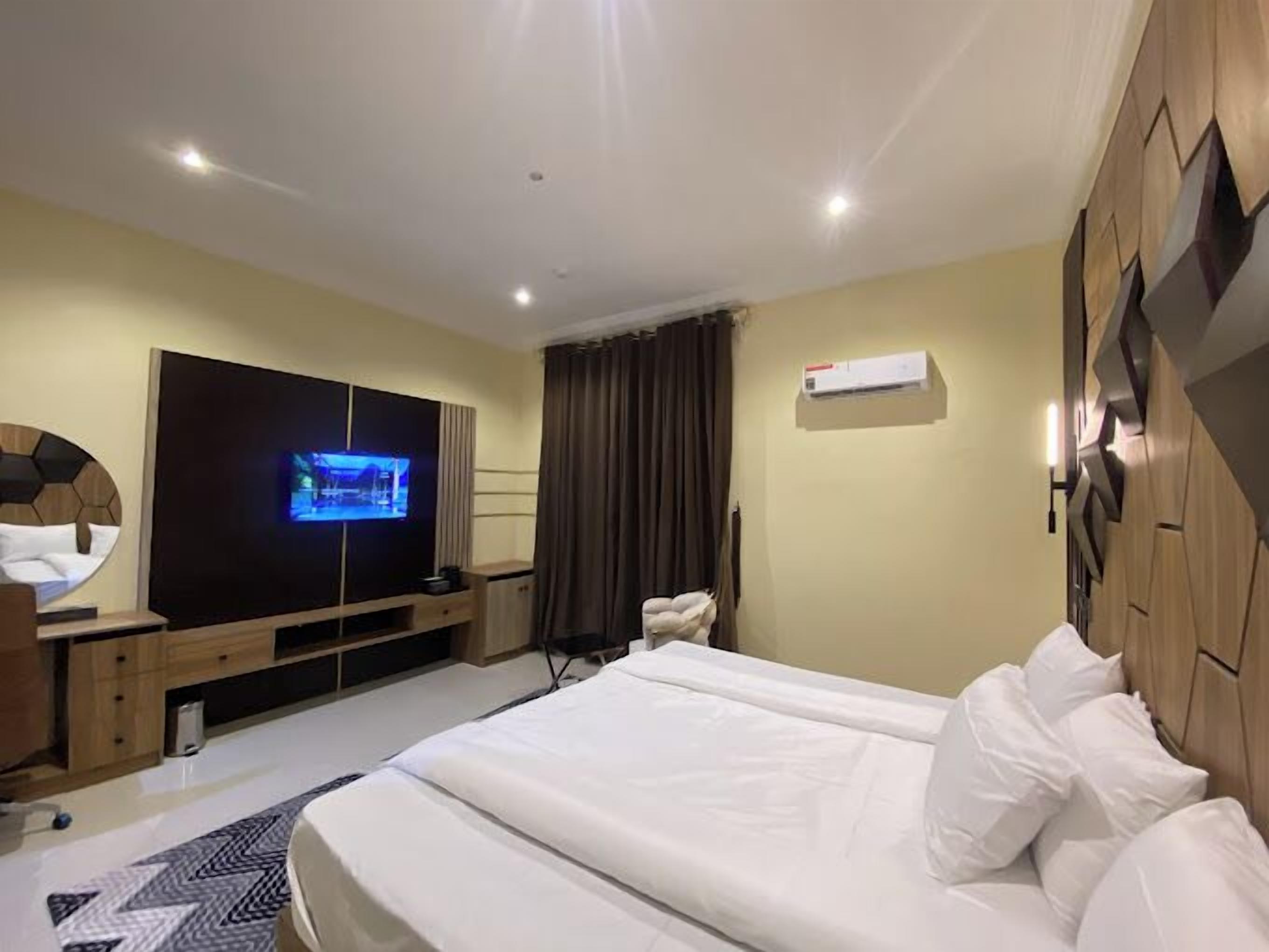 A hotel room with a large bed, a flat-screen TV mounted on the wall, a wooden wardrobe, and a small desk with a chair.