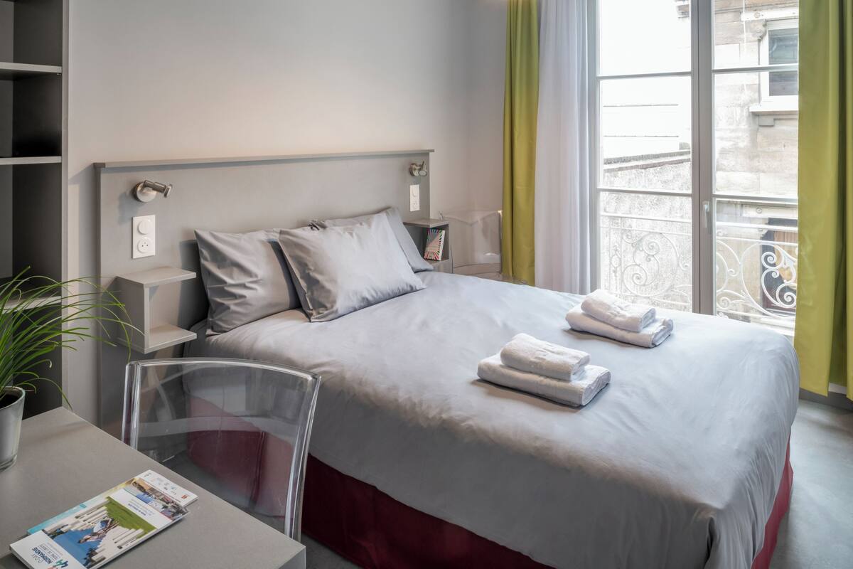 A hotel room with a bed, a desk, a chair, a potted plant, and a window with a balcony view.