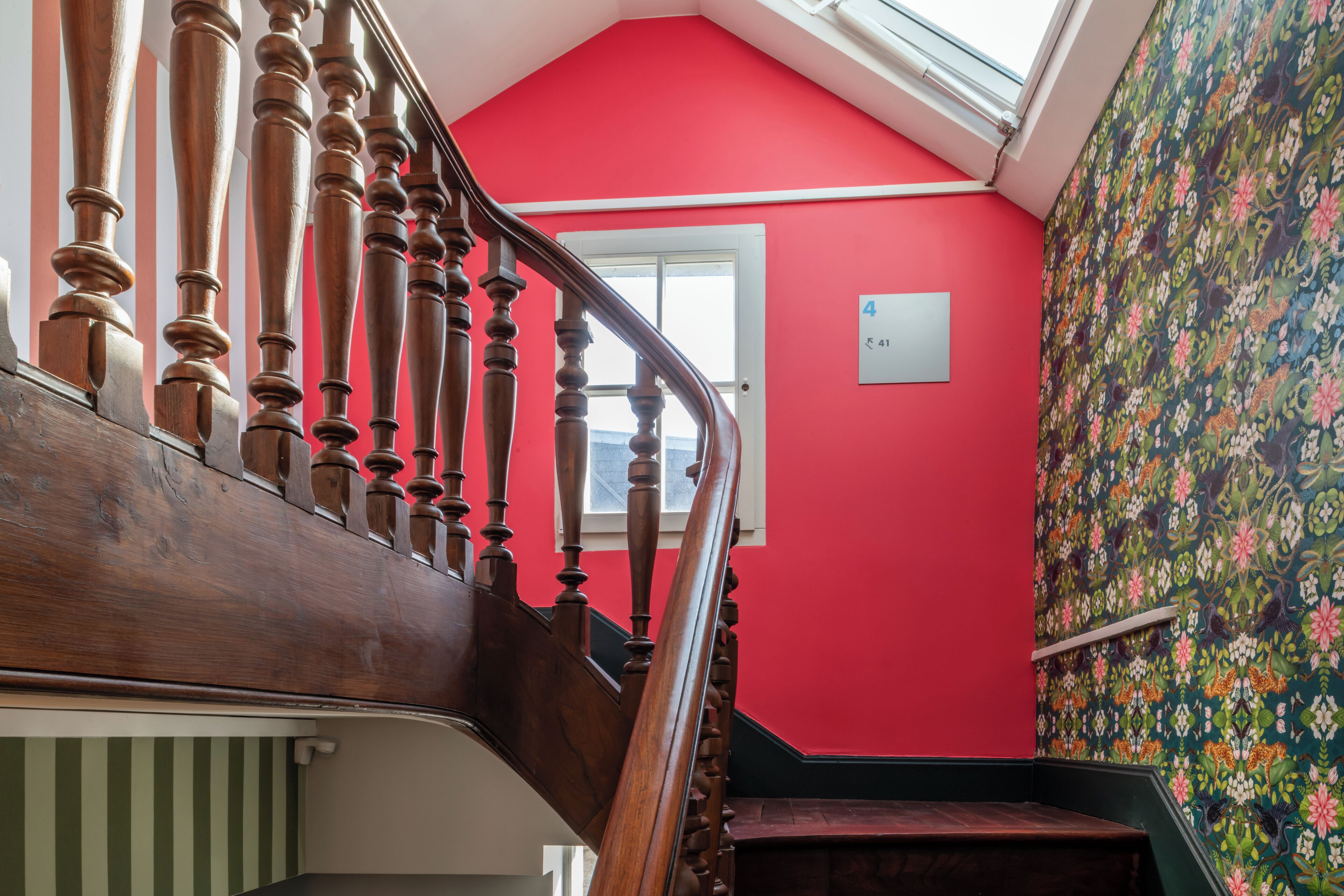 Un escalier avec des balustrades en bois menant à un palier doté d’un mur rouge et d’un papier peint à motifs floraux.