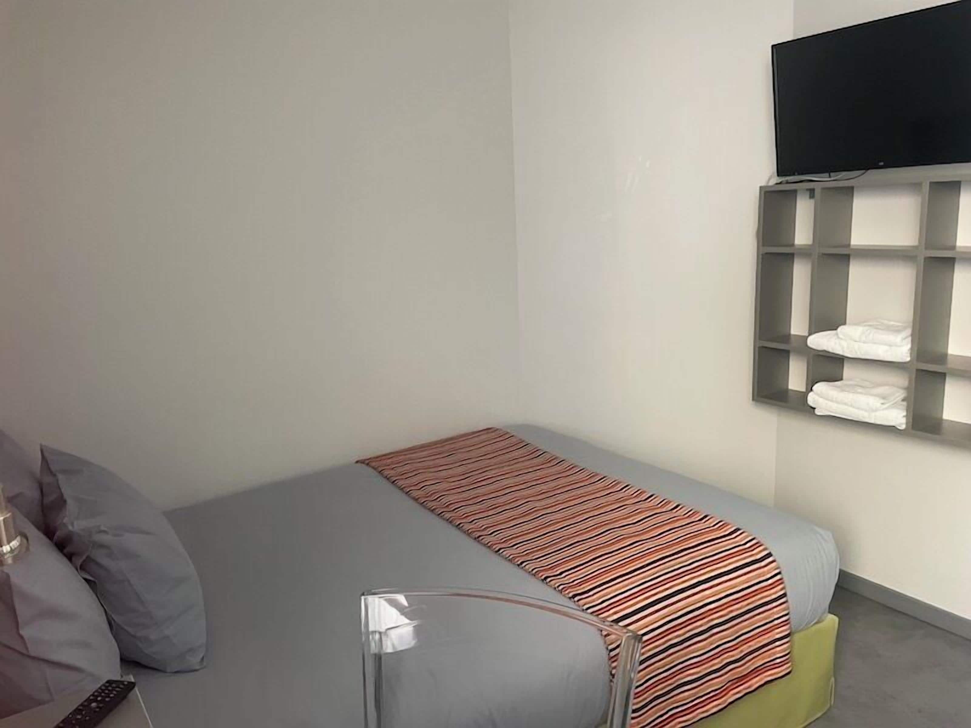 A small, neatly arranged bedroom with a bed, a TV mounted on the wall, and a shelf with folded towels.