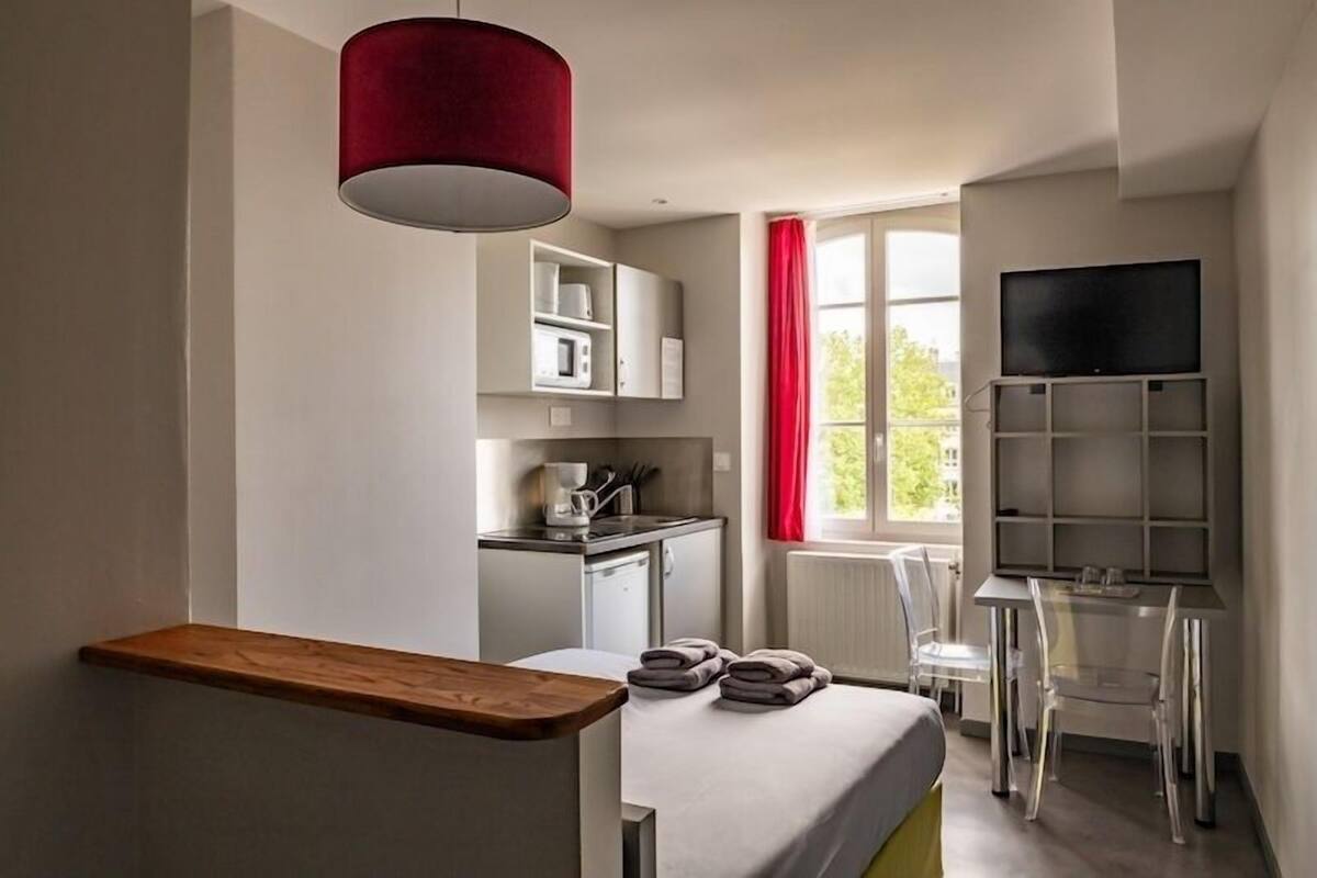 A compact hotel room with a bed, a small kitchenette, a TV, and a window with red curtains.