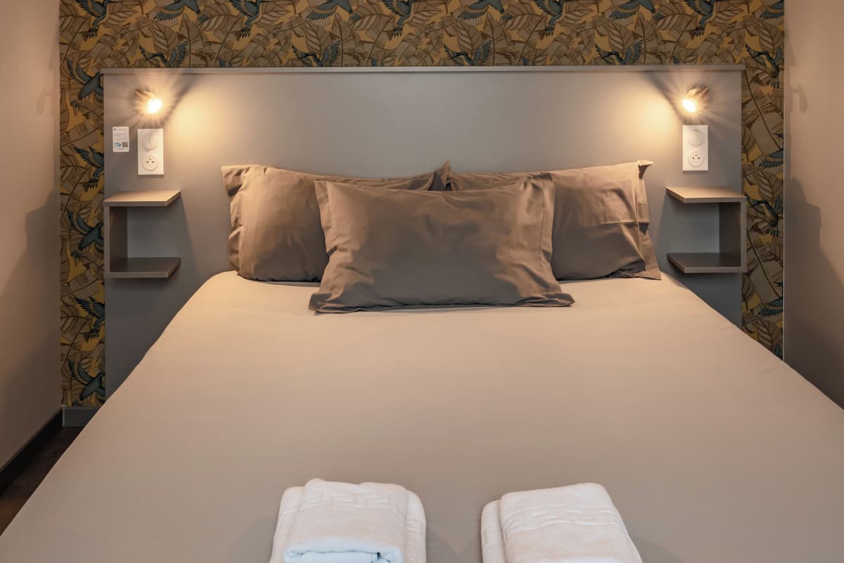 A neatly made bed with two folded white towels, a headboard with a light fixture, and a wall with a leaf-patterned wallpaper.