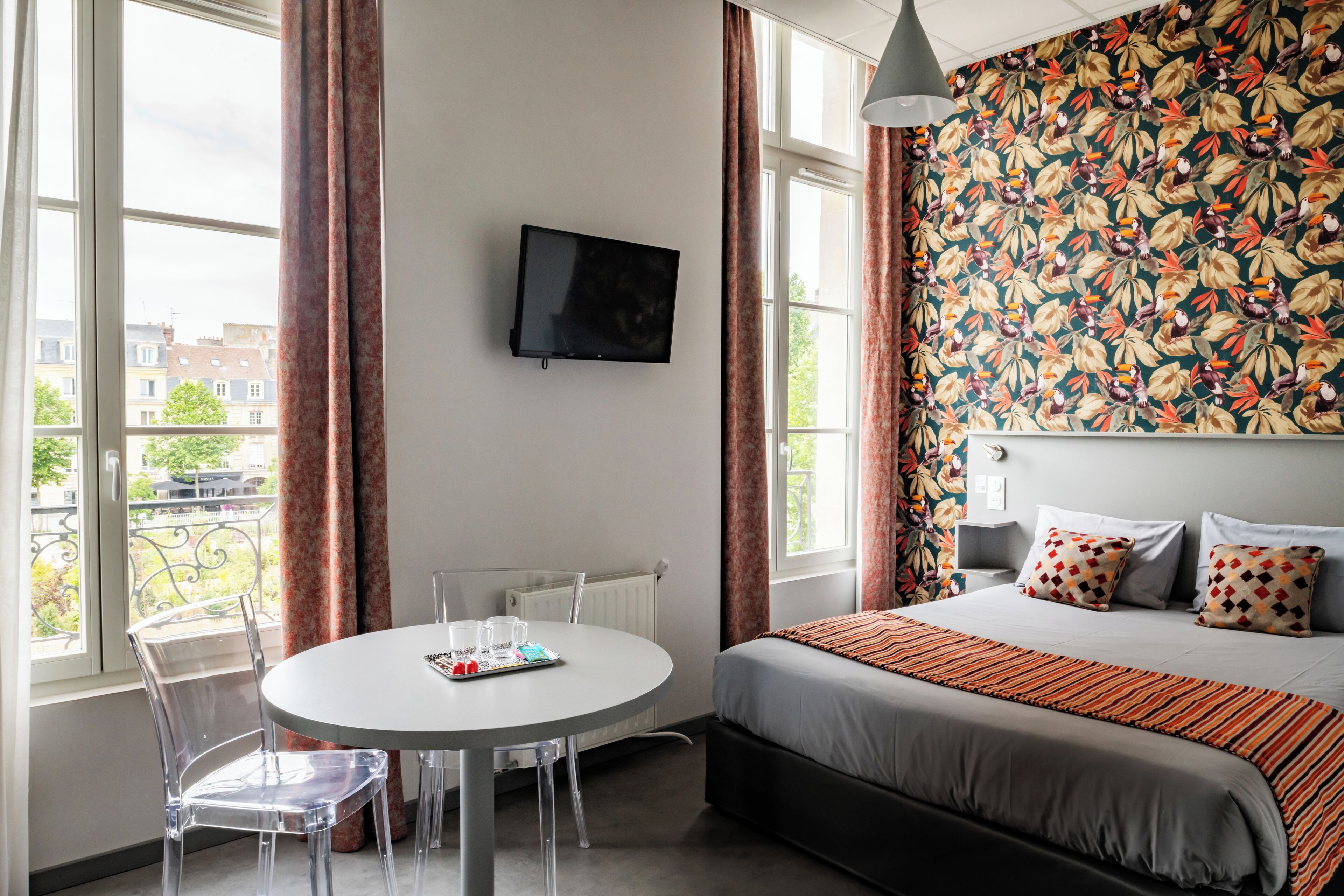 Ein Hotelzimmer mit Bett, einem an der Wand befestigten Fernseher, einem runden Tisch mit Stühlen und einem Fenster mit Vorhängen.