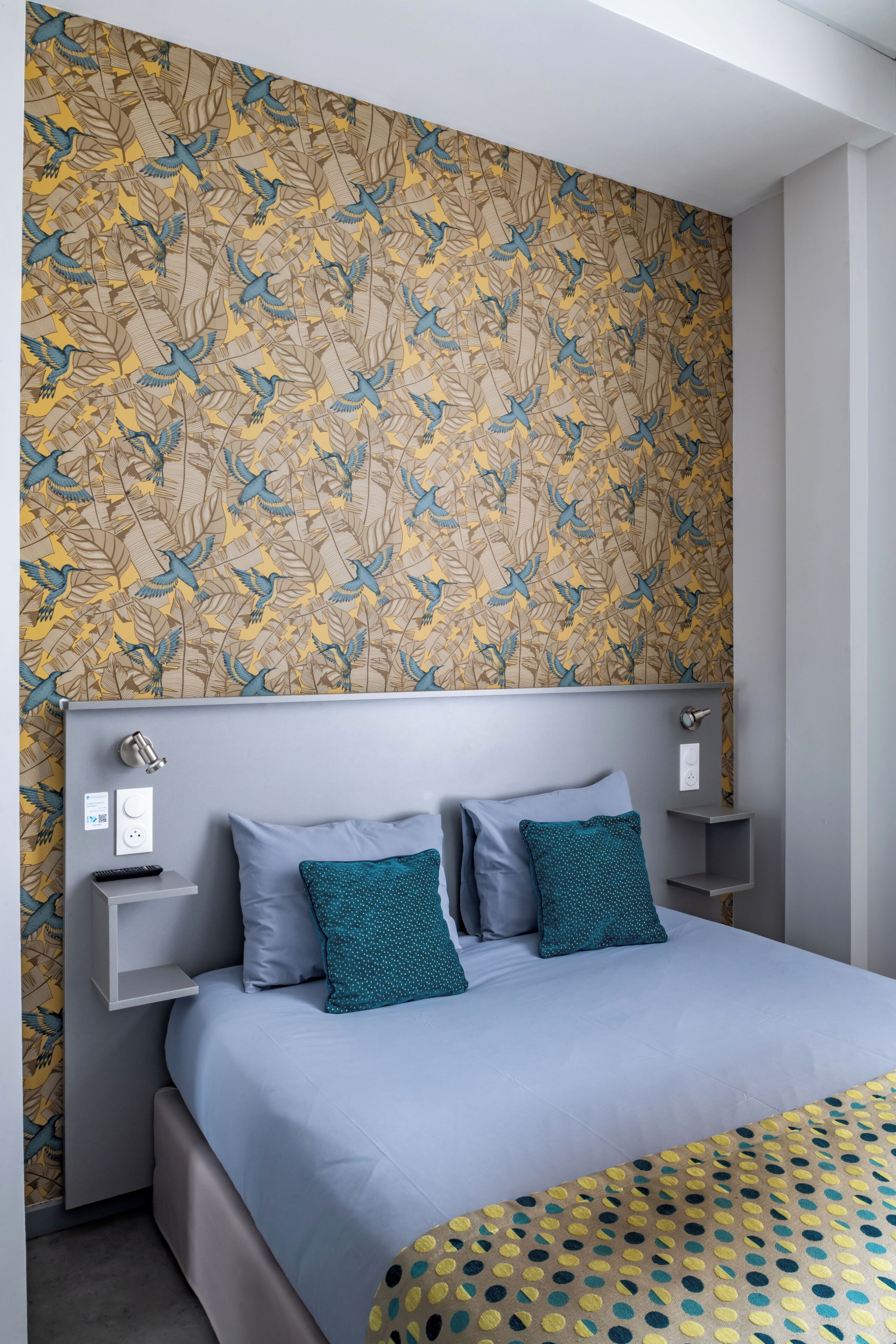 Une chambre à coucher moderne avec un papier peint à motifs, une tête de lit grise et un lit agrémenté d’oreillers bleus et turquoise.