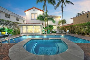 Une piscine avec un jacuzzi, entourée d’une terrasse aménagée avec des meubles d’extérieur et des palmiers.