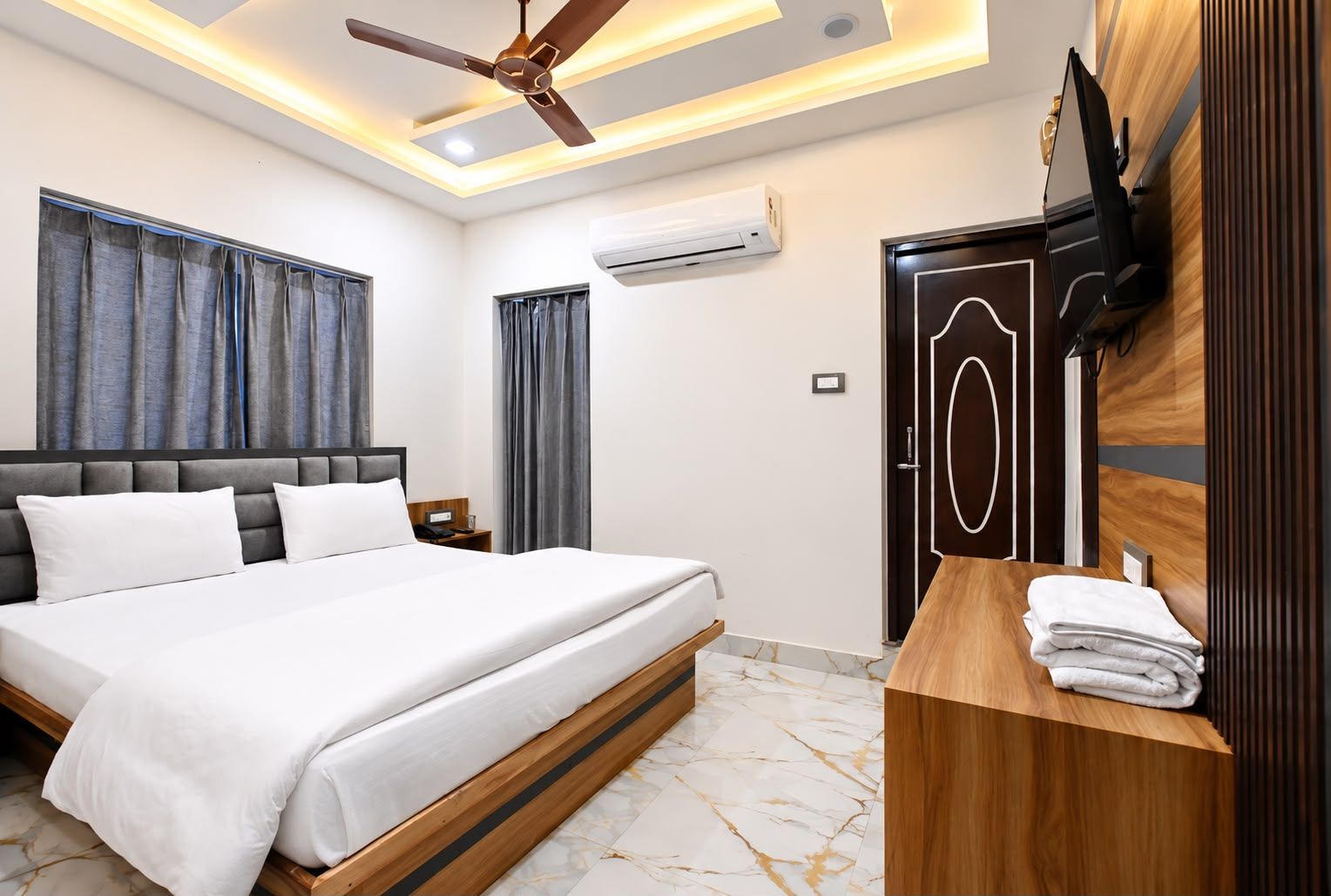 Une chambre d’hôtel comprenant un lit, une table de chevet en bois avec un téléviseur, un ventilateur de plafond et un appareil de climatisation.
