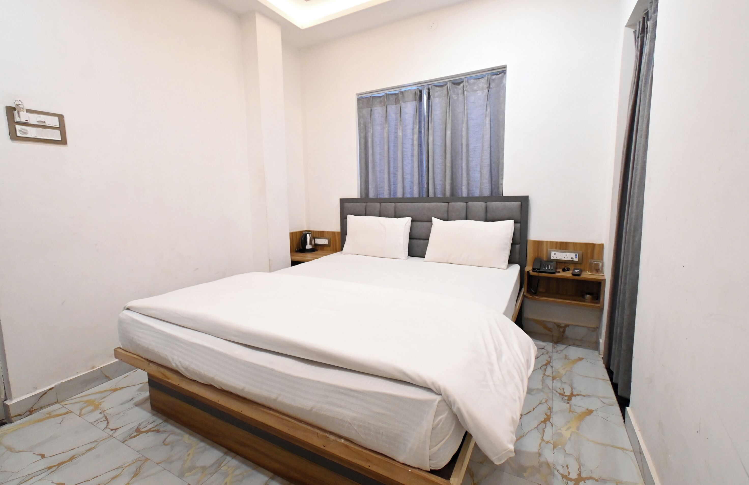 A bedroom with a large bed, a nightstand, a wooden headboard, a window with curtains, and a wall-mounted light fixture.