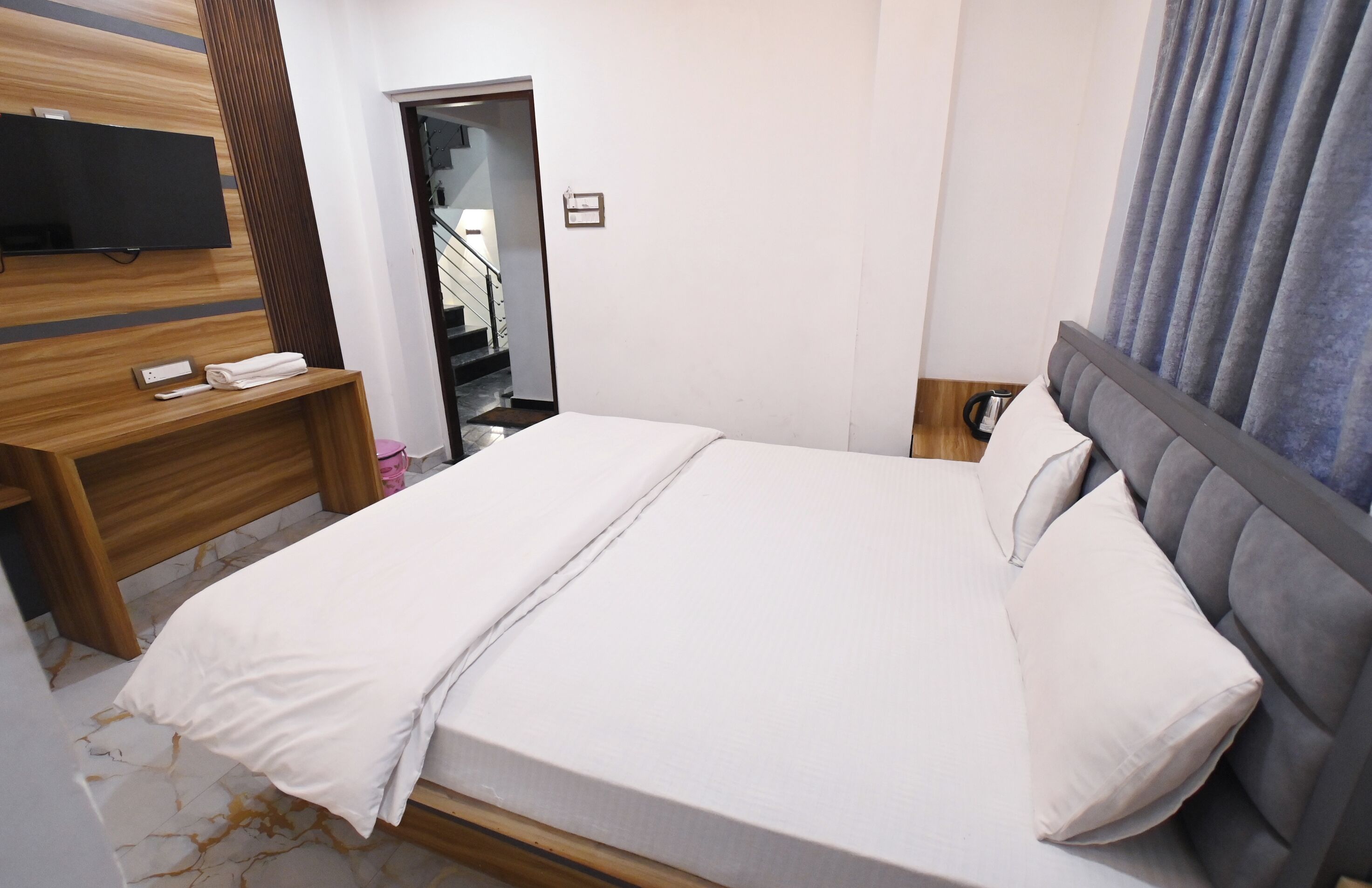 Een net opgemaakt bed met een grijs hoofdbord, een houten nachtkastje met een afstandsbediening, en een flatscreen televisie aan de muur.