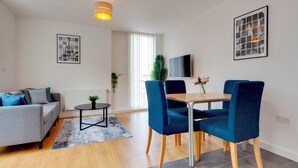 A modern living room with a grey sofa, a small round table, and two blue chairs. There is a TV mounted on the wall and a window with blinds.